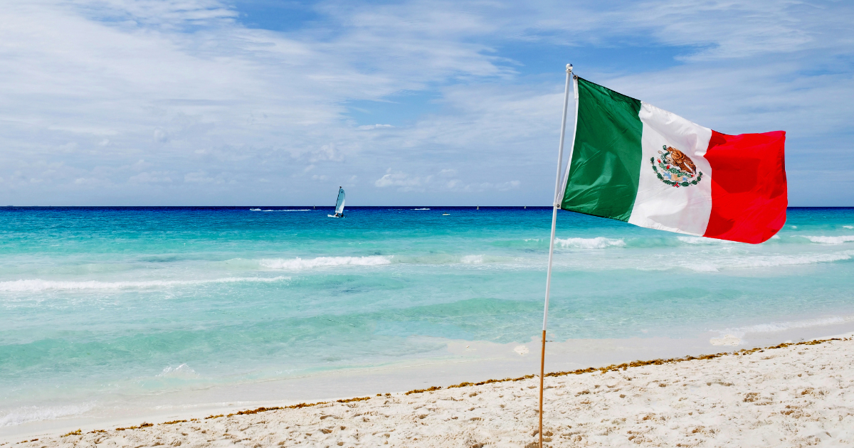 A beautiful Mexican beach with a flag and sailboats on the horizon, under a clear blue sky with fluffy clouds.