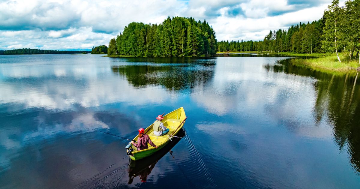 Two people fishing in a yellow rowboat on a tranquil lake surrounded by lush green forests, with a cloudy sky reflecting on the water.