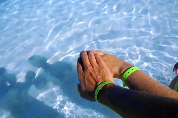 Two hands wearing green wristbands, touching the surface of clear blue water.