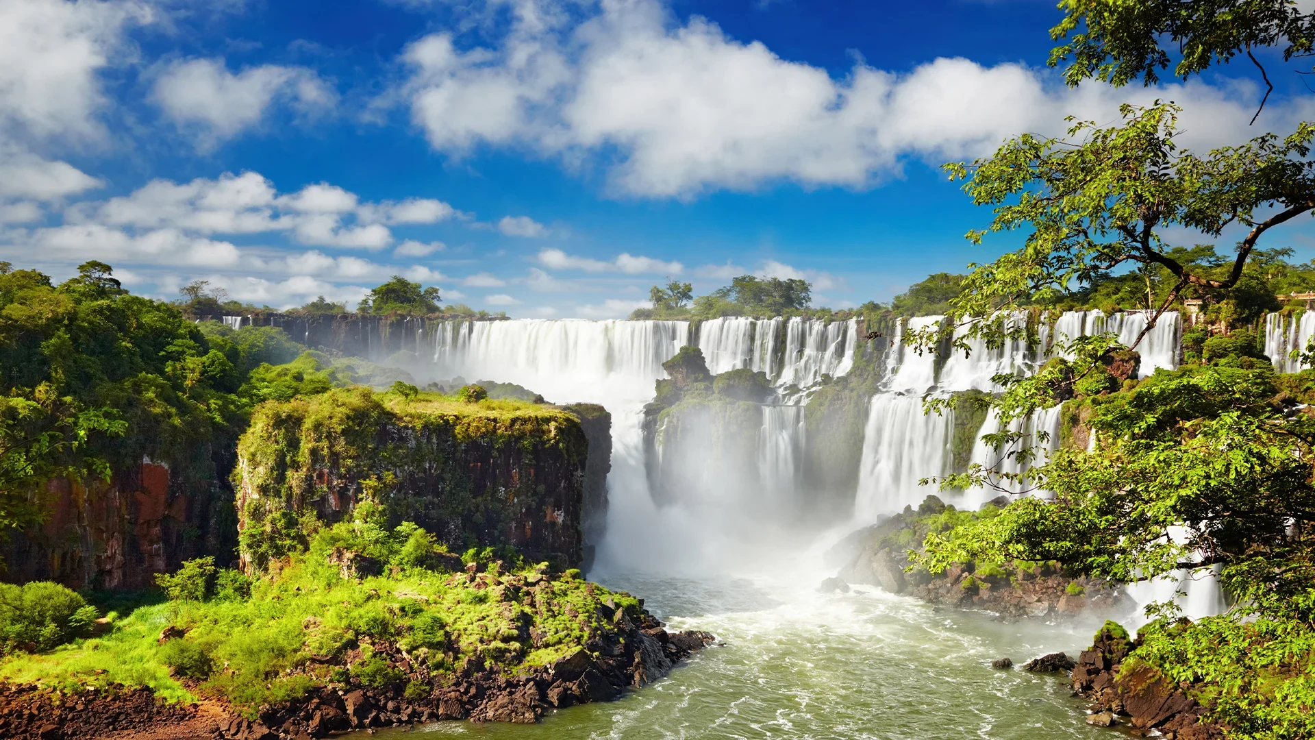 A panoramic view of the Iguazu Falls, with cascading water and lush greenery against a blue sky with clouds.
