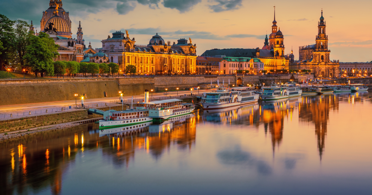 The illuminated skyline of Dresden, Germany, at twilight, with the Elbe River, historic buildings, and boats.