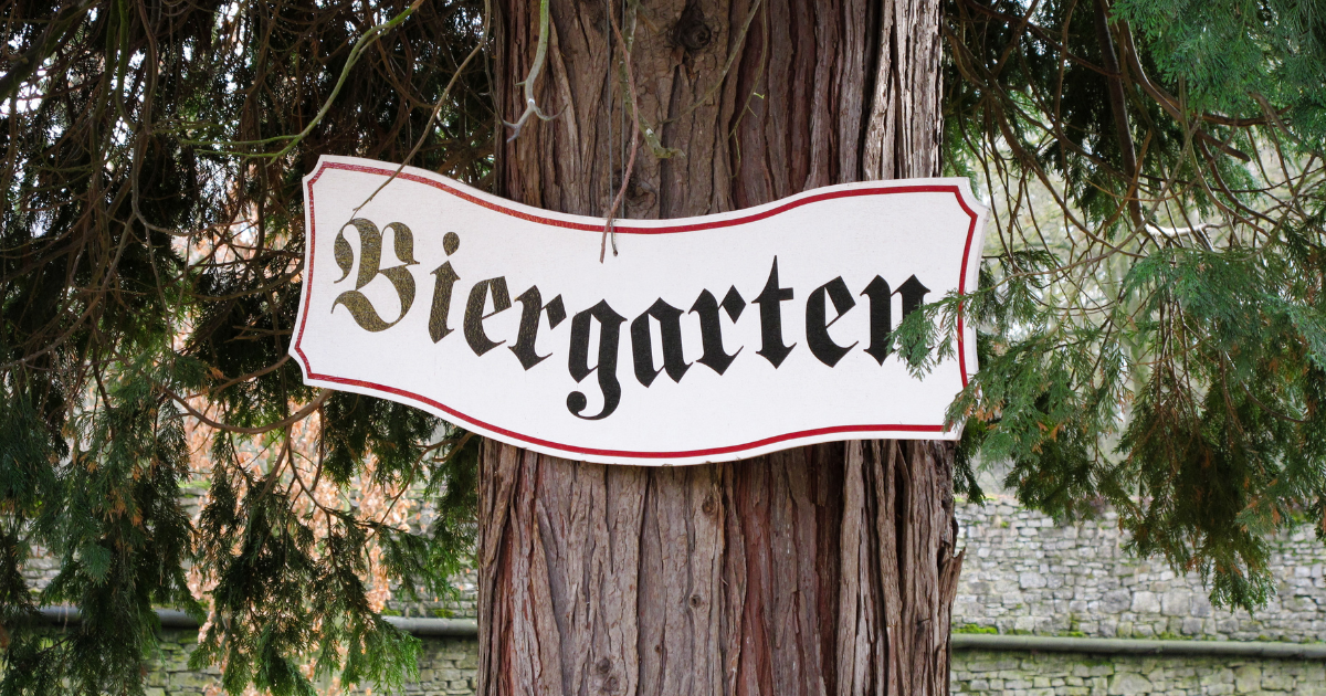 A vintage-style sign with the words "Biergarten" painted in black on a white background, hanging from a tree branch.