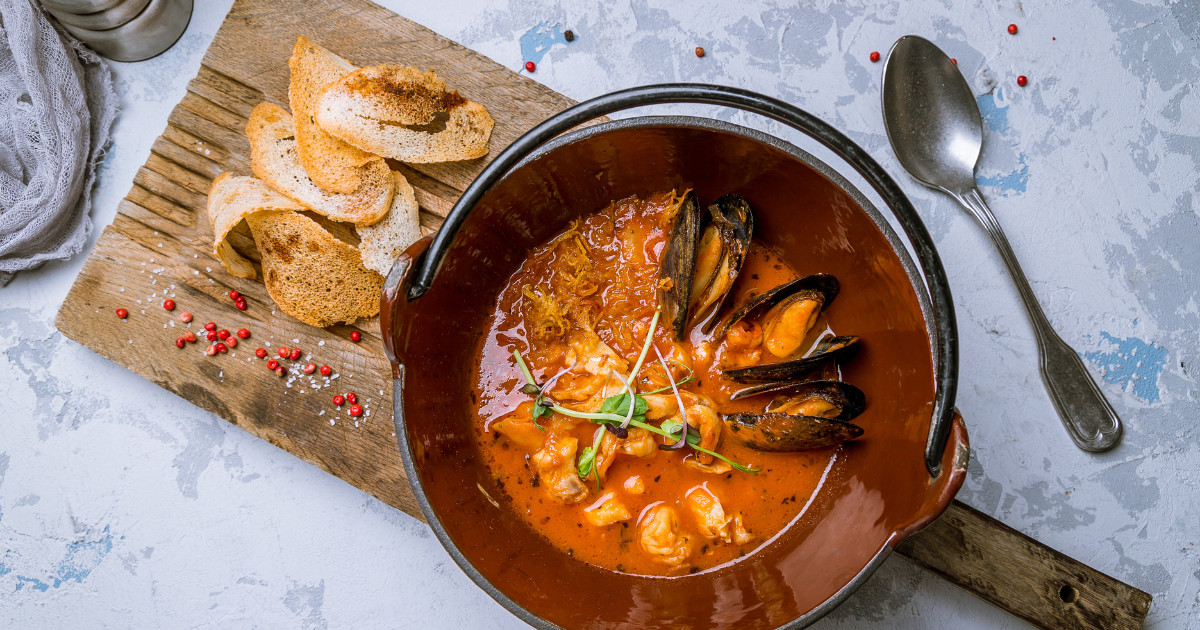 A top-down view of a bowl of seafood bouillabaisse with mussels, fish, vegetables, and croutons, served on a wooden board.