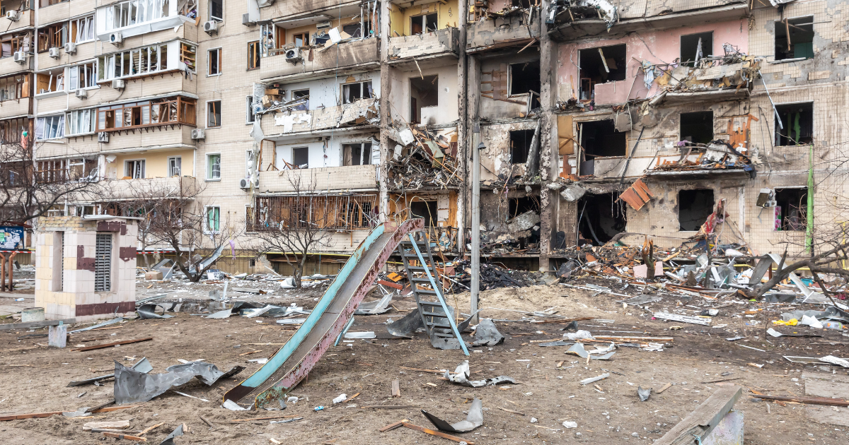 A residential building heavily damaged by war, with shattered windows, collapsed balconies, and debris scattered around a playground.