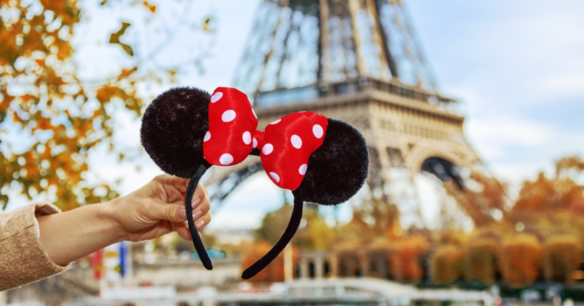 A hand holds up a pair of black Mickey Mouse ears with a red polka-dot bow in front of the Eiffel Tower in Paris.