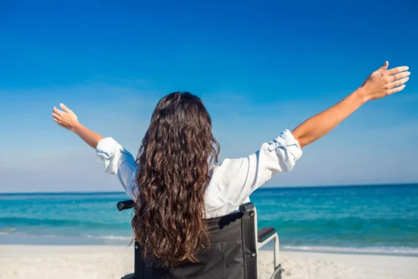 The image shows a woman in a wheelchair with her arms raised, enjoying a sunny day at the beach with a vibrant blue sky and ocean in the background.
