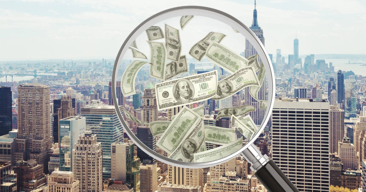 A magnifying glass focusing on US dollar bills, with the New York City skyline visible in the background.