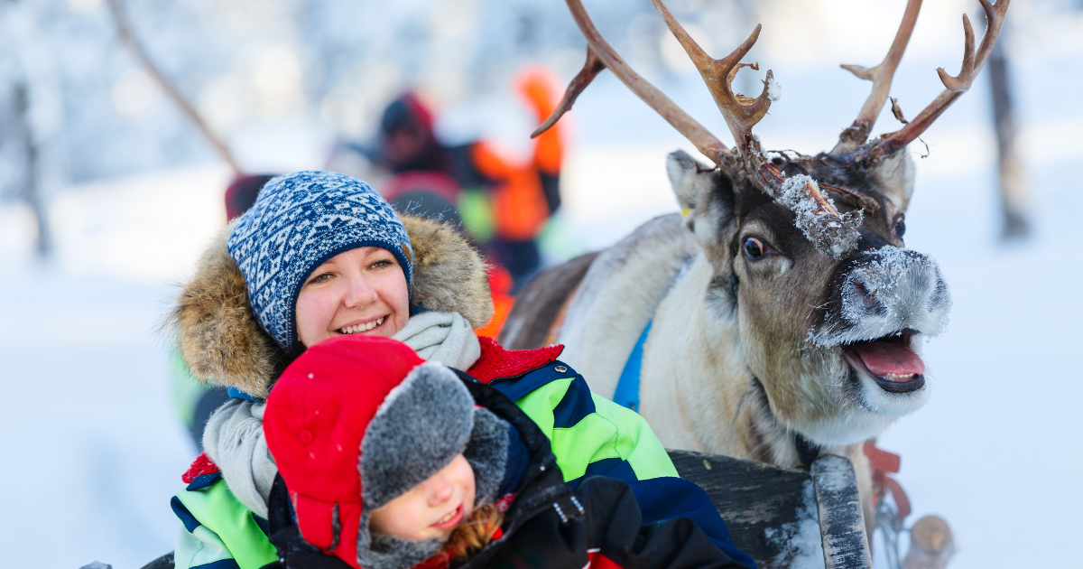 A woman and child smiling on a reindeer sleigh ride in a snowy landscape.