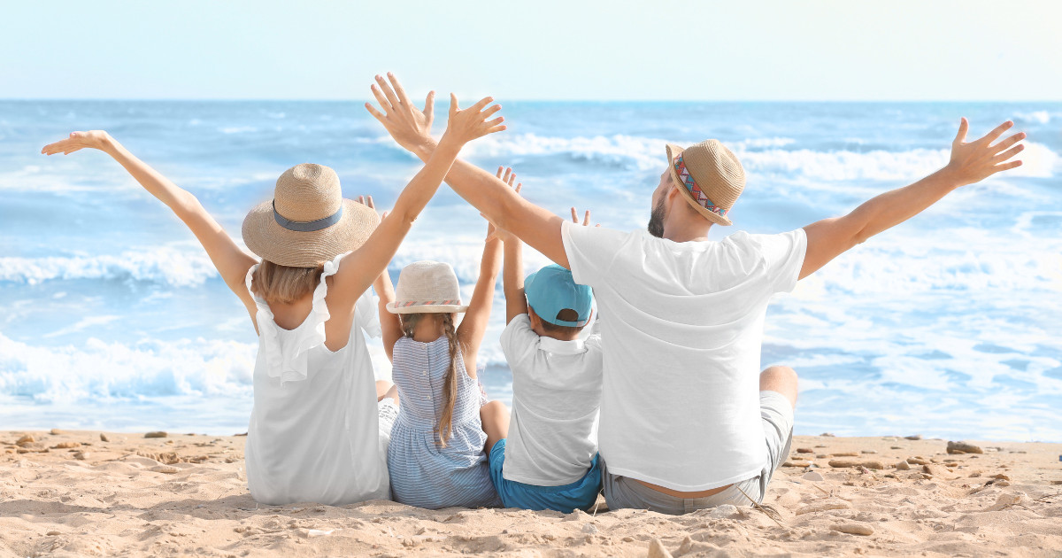 A family of four sits on a beach with their backs to the camera, all raising their arms in joy. They wear summer clothes and hats while facing the ocean waves.