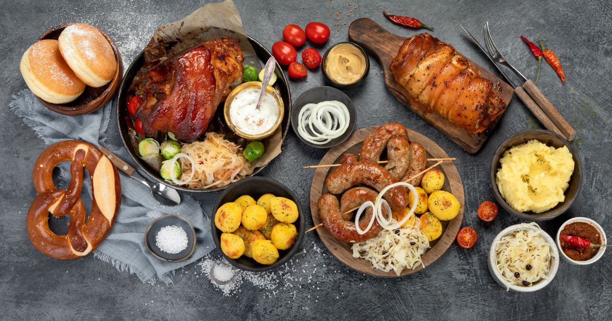 A delicious spread of German food, featuring roasted pork knuckle, sausages, grilled potatoes, sauerkraut, pretzels, and various dipping sauces.