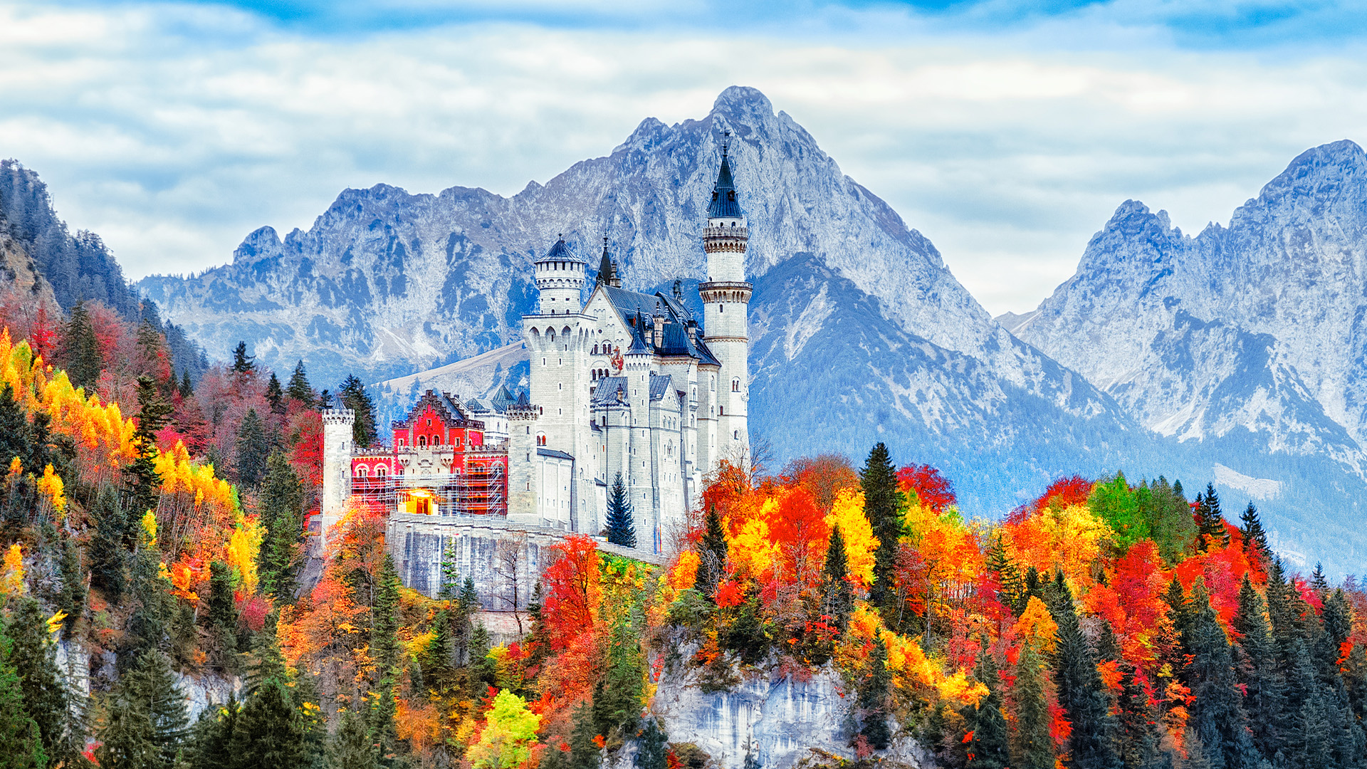 A breathtaking view of Neuschwanstein Castle, a 19th-century Romanesque Revival palace perched on a hilltop in Bavaria, Germany, with a backdrop of snow-capped mountains and autumn foliage.