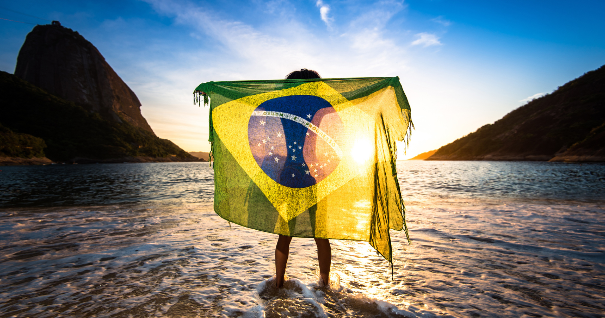 A person standing in the water, holding the Brazilian flag in front of them, with Sugarloaf Mountain in the background.