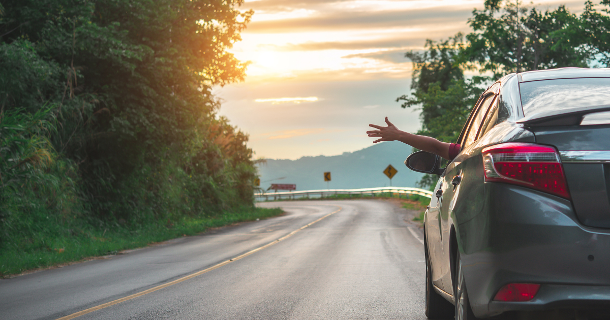 A person waving their hand out of a car window, enjoying a scenic road trip with a sunset in the background.