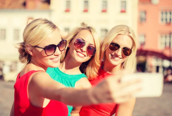 The image shows three young women wearing sunglasses and colorful clothes, standing together and smiling in an urban setting.