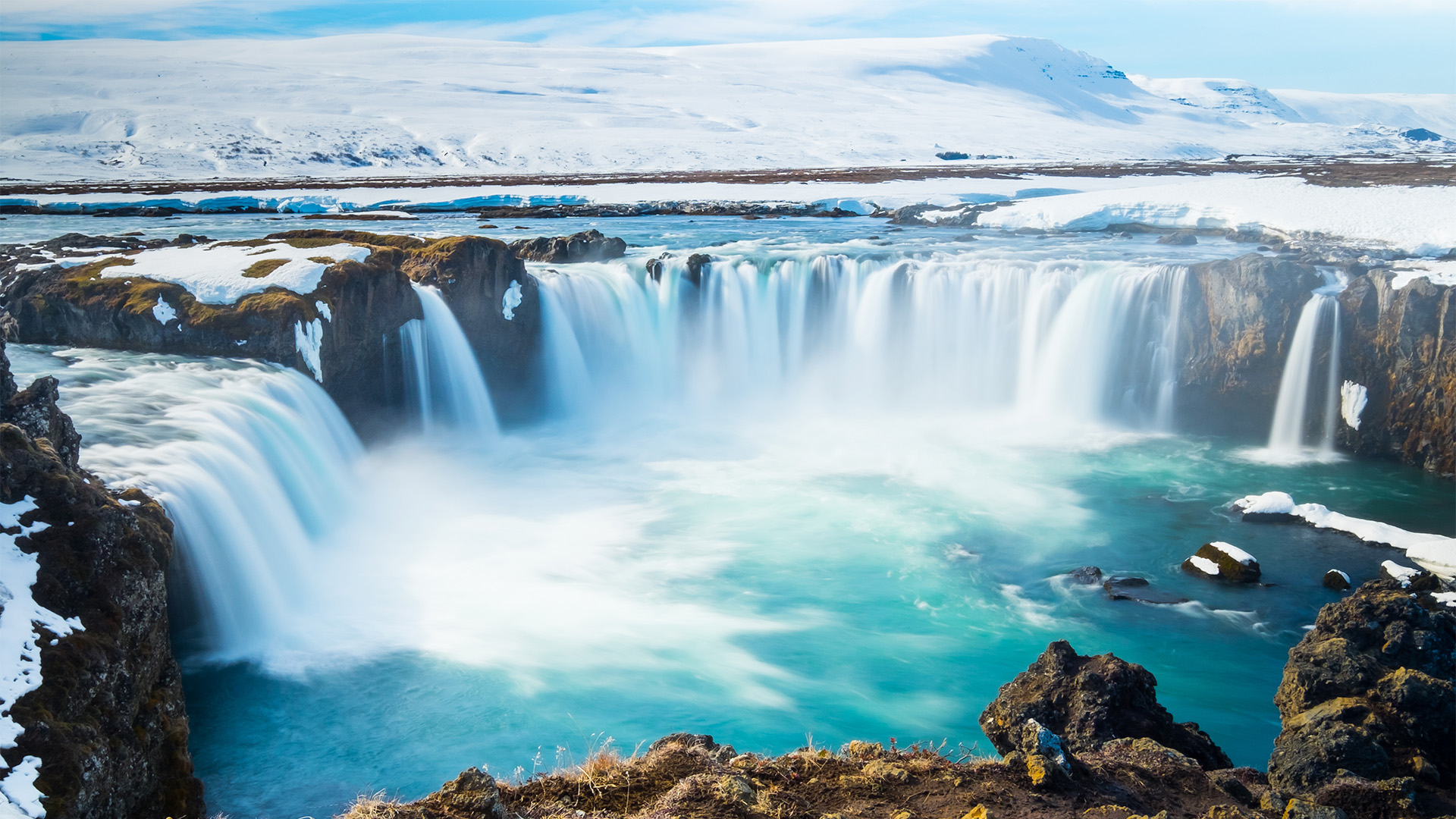 A breathtaking view of the cascading Dettifoss waterfall in Iceland, with snow-capped mountains and a vibrant turquoise pool in the foreground.