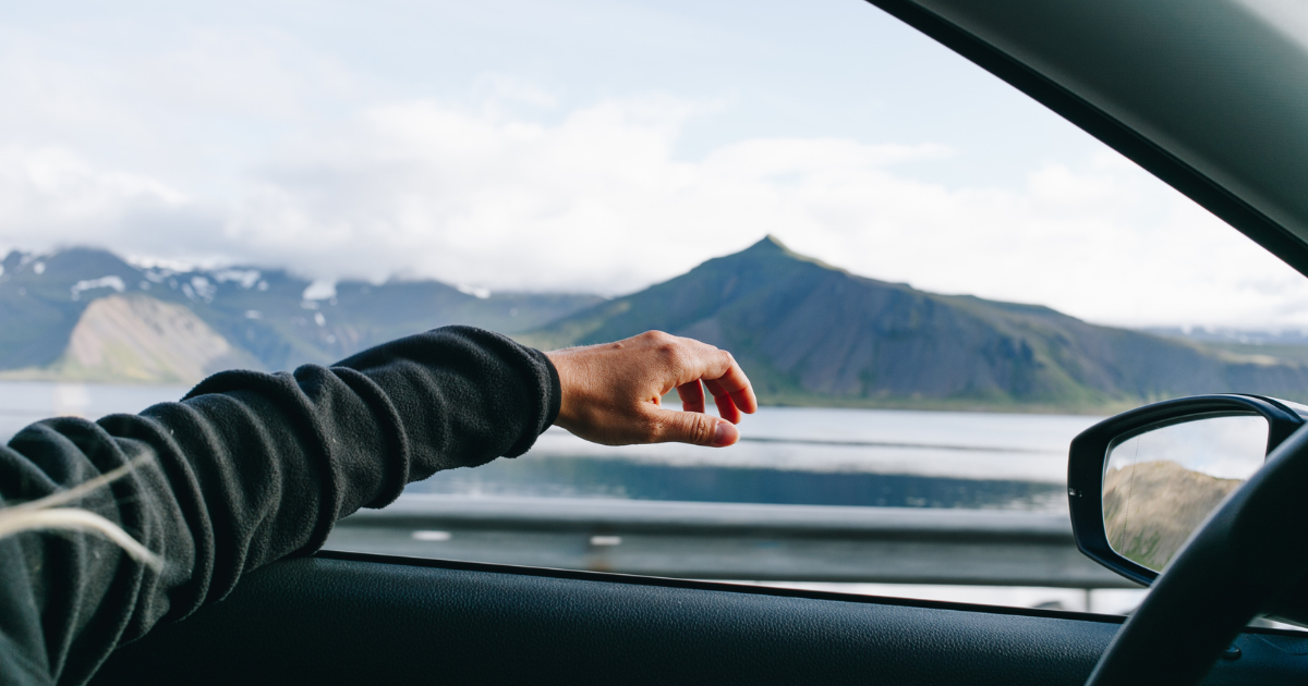 A person's hand reaching out of a car window with a scenic mountain view.