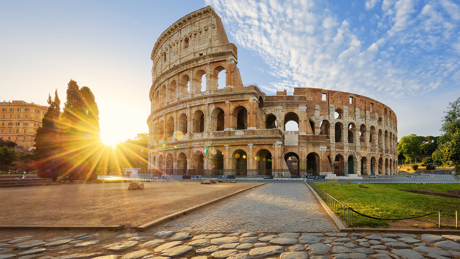 The iconic Colosseum in Rome, bathed in the warm light of sunrise, with a clear blue sky and sunbeams.