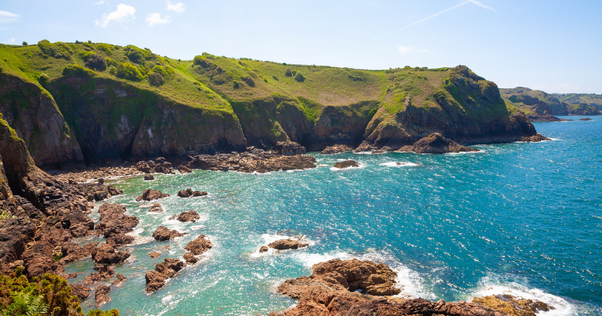 A breathtaking view of the rugged coastline of Jersey, with green cliffs, rocky shores, and a sparkling blue sea.