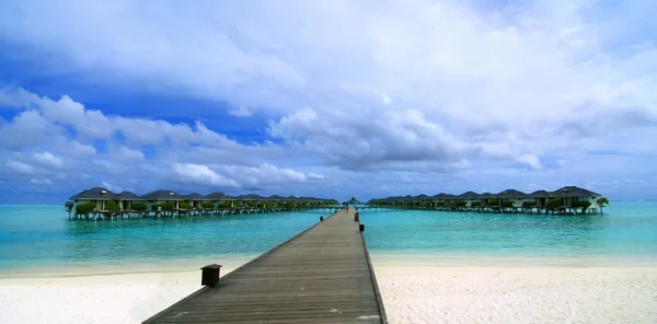 A wooden walkway leading to overwater bungalows with a view of the turquoise ocean and white sandy beach.