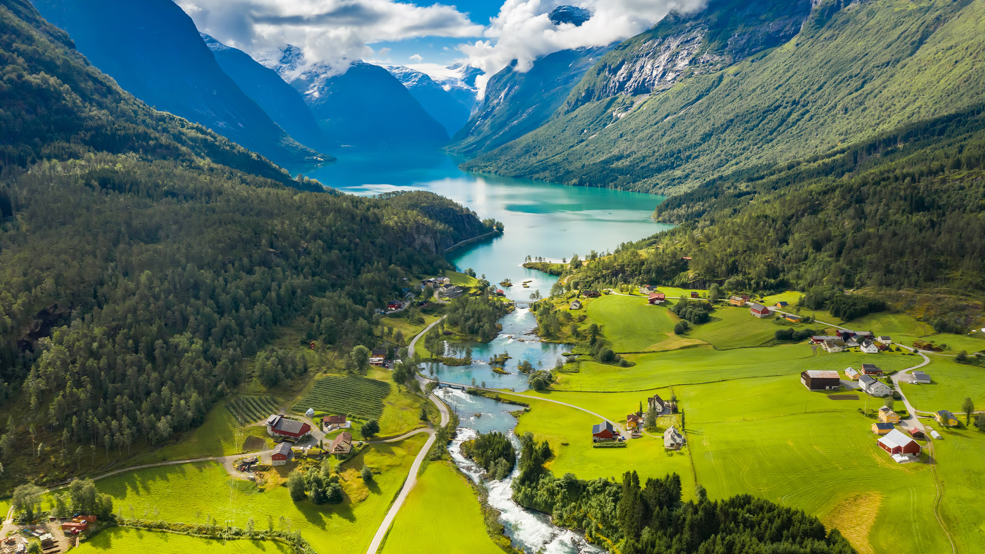 A breathtaking aerial view of a picturesque valley in Norway, showcasing a crystal-clear lake surrounded by lush green fields, forests, and majestic snow-capped mountains.