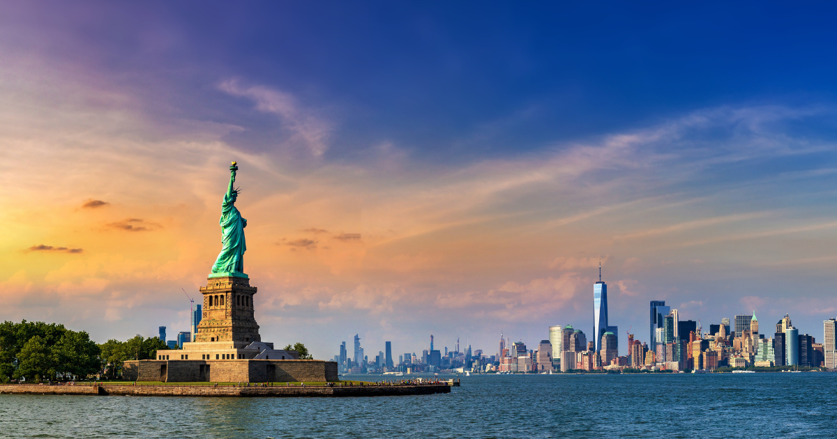 The iconic Statue of Liberty standing tall on Liberty Island, with the stunning New York City skyline in the background, at sunset.