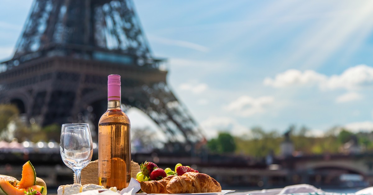 A picnic spread with wine, cheese, fruit, and pastries, with the Eiffel Tower in the background.