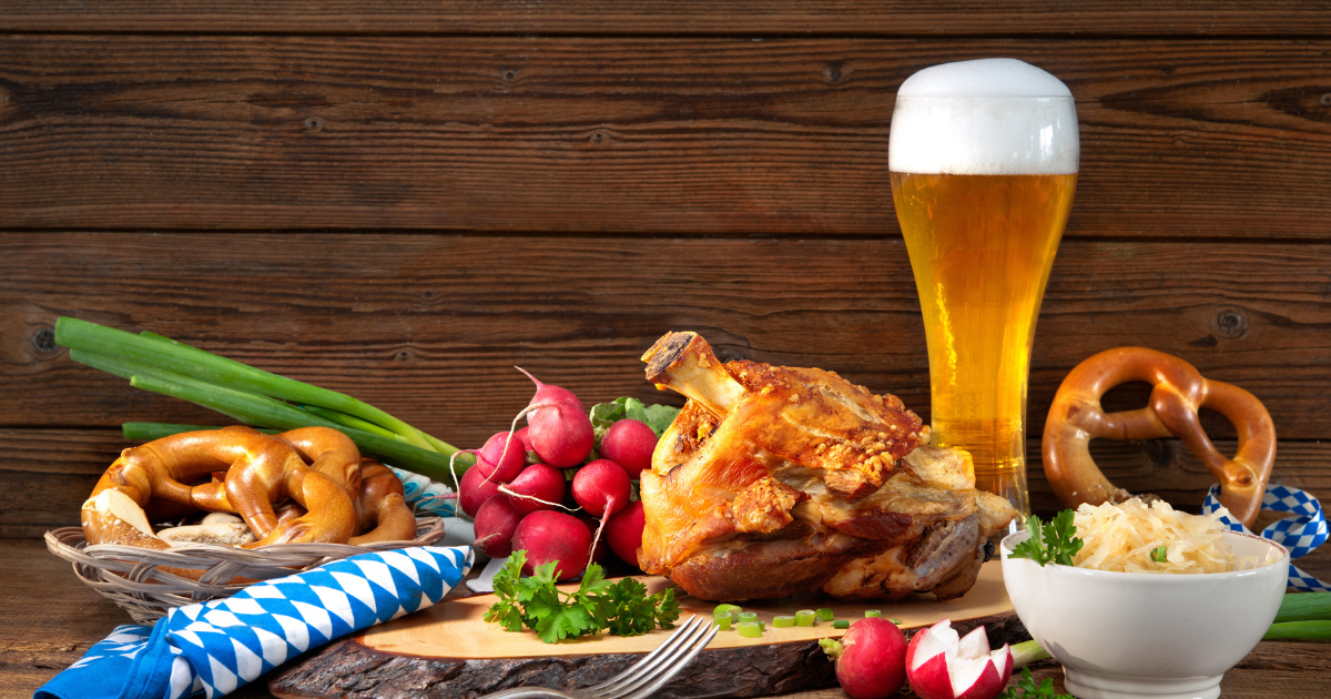 A festive Oktoberfest spread featuring a crispy roasted pork knuckle, pretzels, radishes, sauerkraut, and a tall glass of beer.