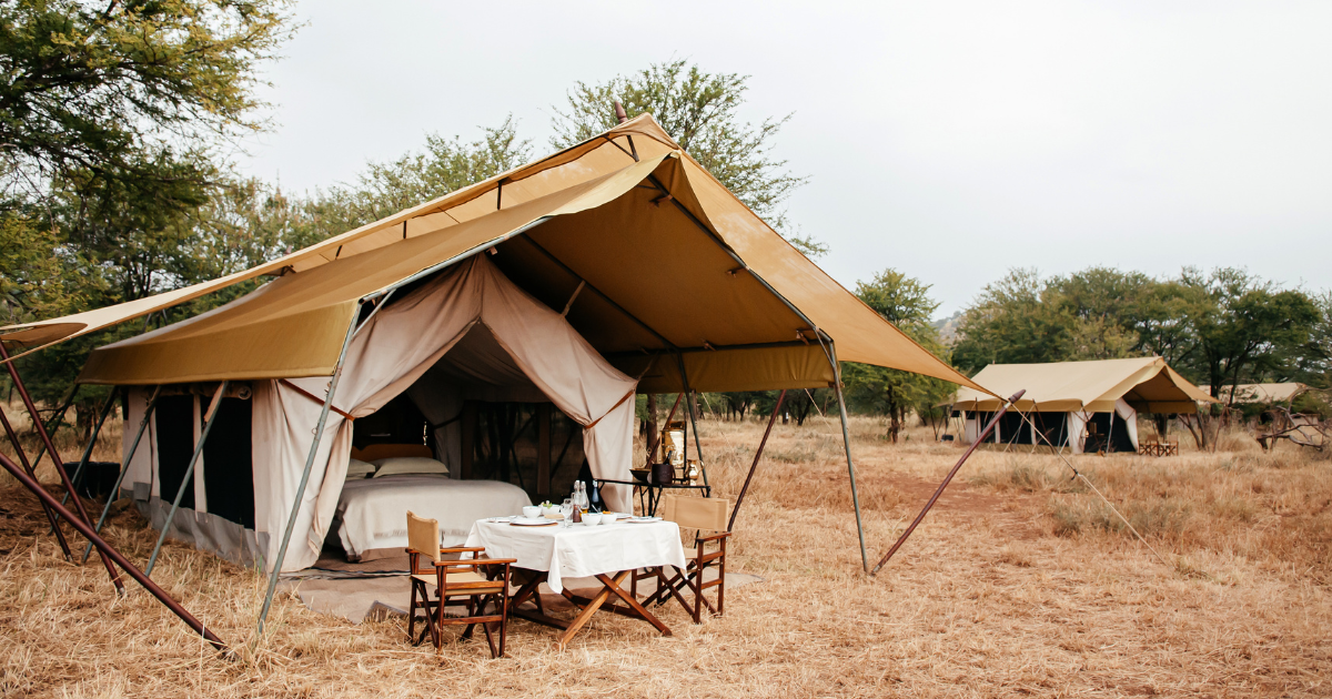 A luxury safari tent camp with comfortable beds, a dining table, and chairs, set in a scenic African savanna.