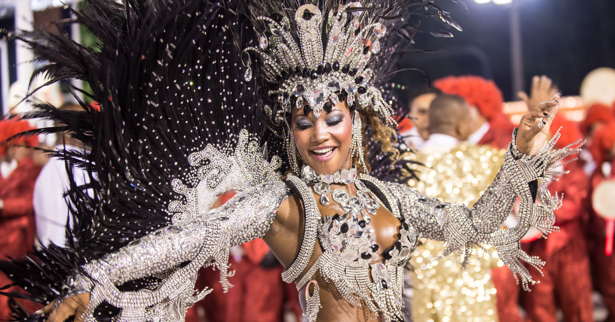 This image shows a performer wearing an elaborate costume decorated with rhinestones and feathers, smiling and gesturing during what appears to be a colorful, festive event or parade.