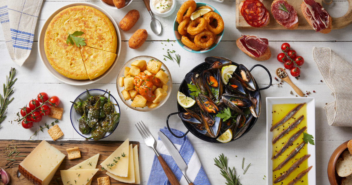 A top-down view of a table filled with a variety of Spanish tapas, such as paella, croquettes, mussels, olives, chorizo, and cheese.