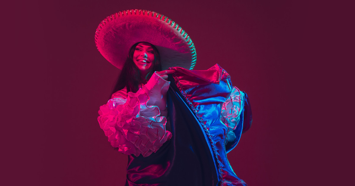 A smiling woman wearing a wide-brimmed Mexican sombrero and a ruffled dress poses against a dark background. The image is lit with dramatic pink and blue neon-style lighting.