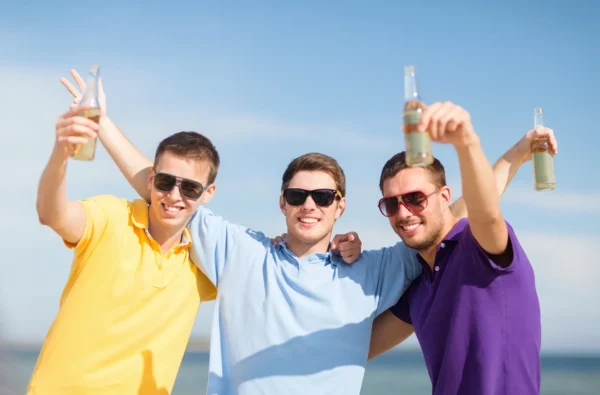 The image shows three young men standing outside, wearing sunglasses and holding drinks, and smiling happily.