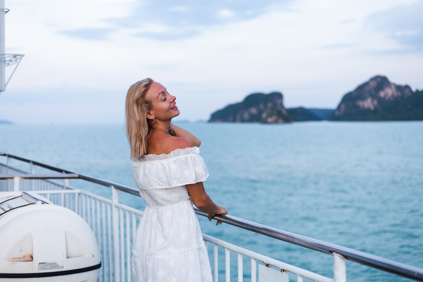 Smiling woman in a white dress enjoying the ocean view from a cruise deck.