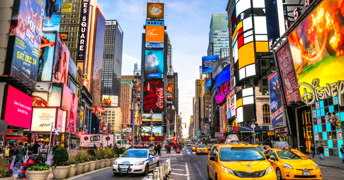 A vibrant and bustling Times Square, with its iconic billboards, streetlights, and yellow taxis, under a clear blue sky.