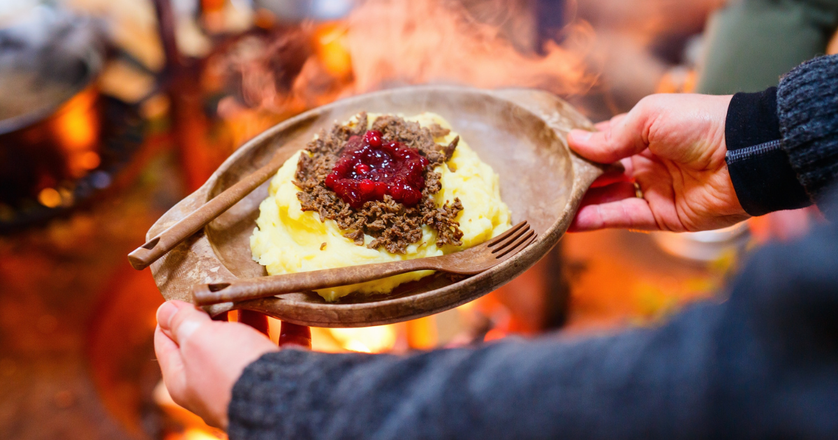 A plate of traditional Finnish food, featuring mashed potatoes topped with a meat sauce and lingonberry jam, held over a campfire.