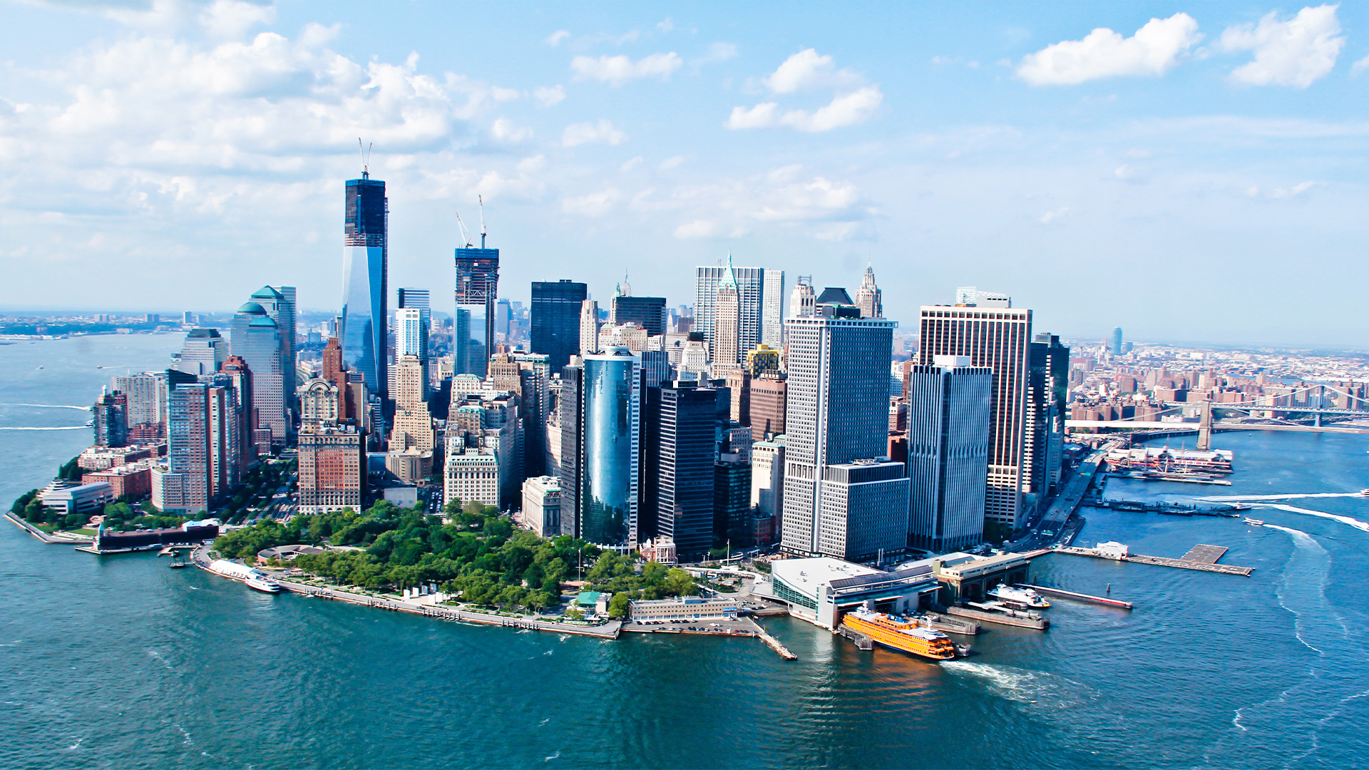 A breathtaking aerial view of Lower Manhattan, New York City, showcasing the iconic skyline with skyscrapers like the One World Trade Center, and the Brooklyn Bridge spanning the East River.