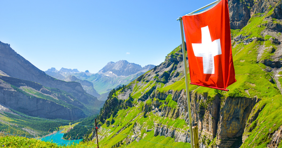 The Swiss flag waving proudly against a backdrop of the Swiss Alps, with a clear blue sky and lush green valleys.