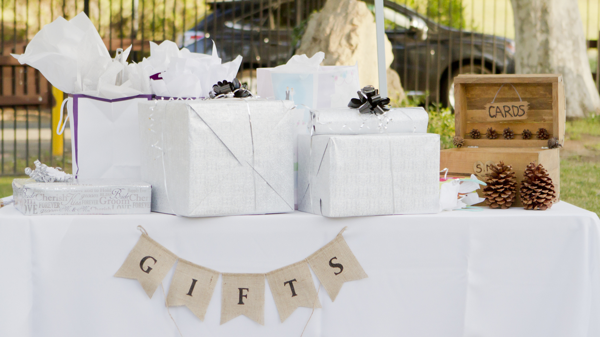 A white table with wrapped wedding gifts, a "Gifts" banner, and a wooden box for cards, set up outdoors.