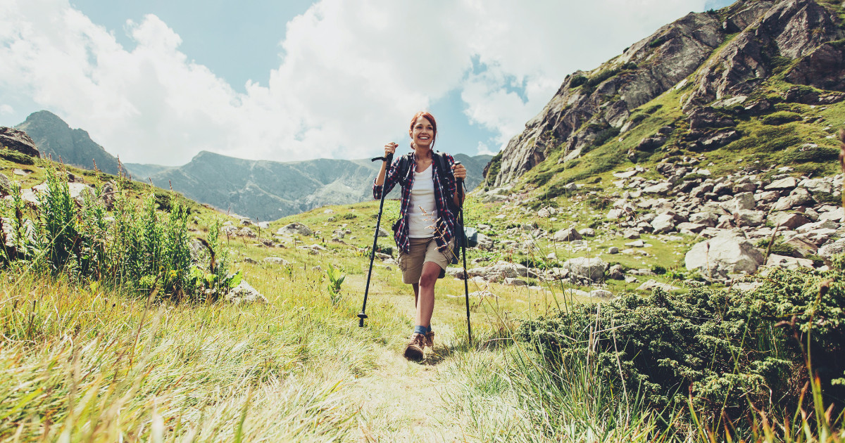 A smiling hiker with hiking poles walks on a mountain trail, wearing shorts and a backpack. The scene shows green grass, rocks, and mountain peaks in the background under a partly cloudy sky.