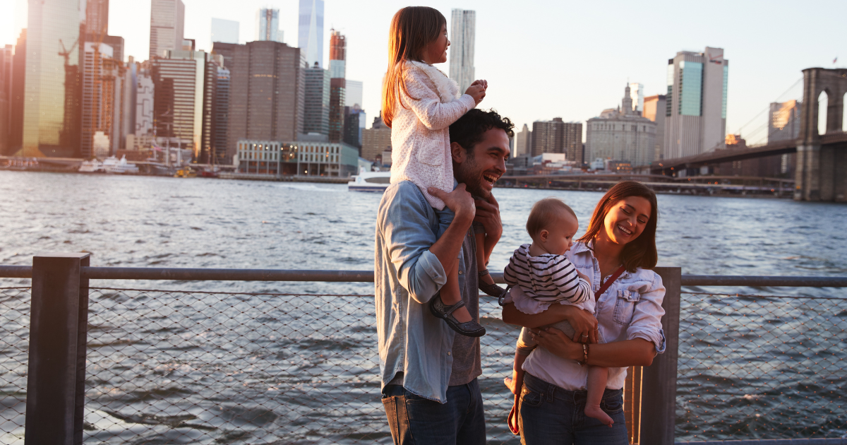 A man is carrying a young child on his shoulders, while a woman stands beside them, holding another child standing by the waterfront, with the New York City skyline in the background.