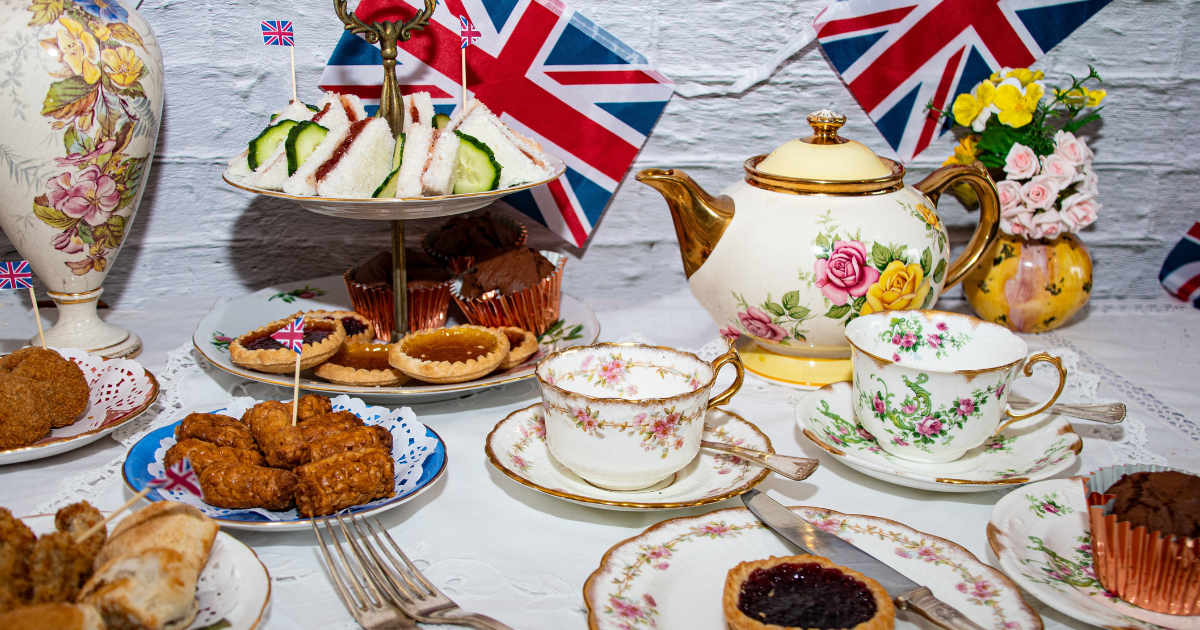 A vintage-style afternoon tea spread with a tiered cake stand, teacups, pastries, sandwiches, and scones, decorated with Union Jack flags and flowers.