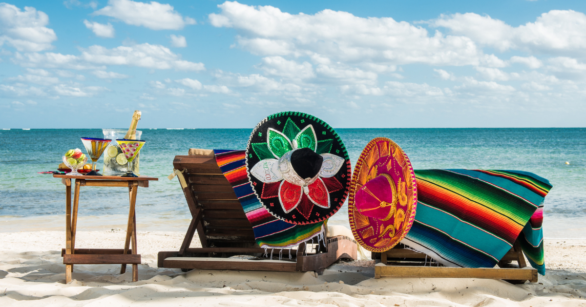 Two beach chairs with colorful Mexican blankets and sombreros, a cocktail table with drinks, on a white sandy beach with a clear blue ocean.