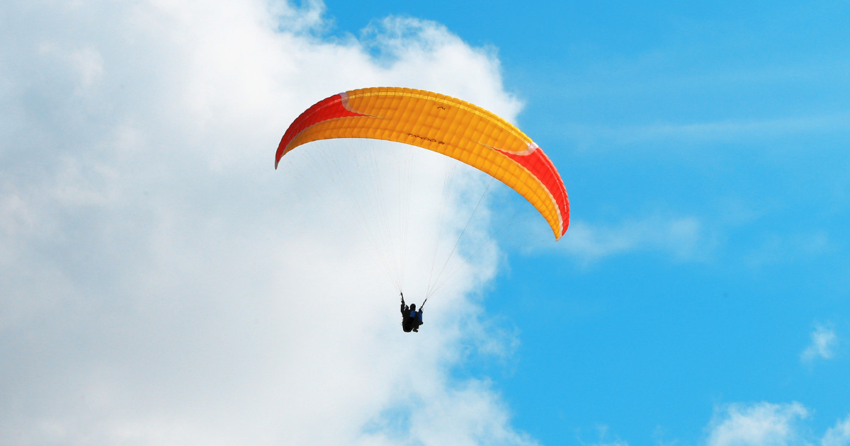 A paraglider with an orange and red canopy soaring through the sky against a backdrop of fluffy white clouds.